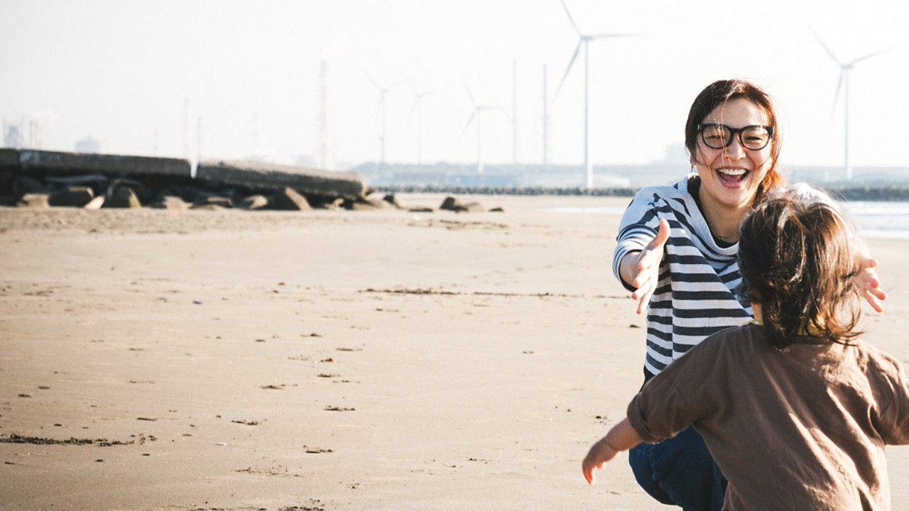 Bērns skrien pie mātes pludmalē ar vējdzirnaviņām